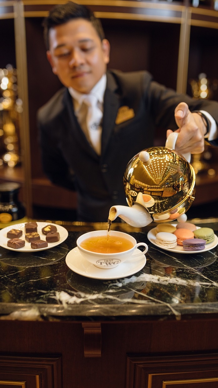 Man pouring tea into a teacup, placed alongside plates of macaroons and chocolates